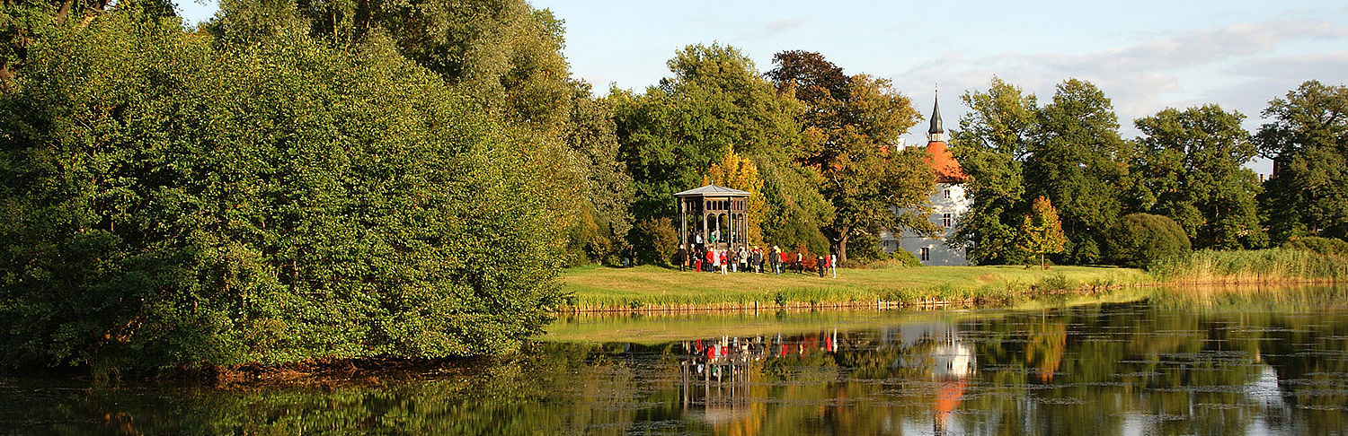 Landschaftspark mit Wasserschloss Fürstlich Drehna © Claudia Donat Bäume, ein Pavillon, ein Schlossturm und eine bunte Schar Parkbesucher spiegeln sich im Parkteich.