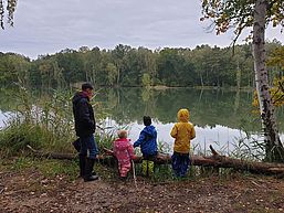Eine Frau und drei kleine Kinder stehen am Ufer eines Sees und schauen auf die glatte Wasseroberfläche.