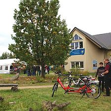 Am Haus des Infozentrums hängt ein Banner mit der Aufschrift "20 Jahre Naturpark Niederlausitzer Landrücken". Im Vordergrund betrachten Leute Fahrräder - darunter ein rotes Tandem. In der Nähe eine Festzeltes sind Festgäste zu sehen.