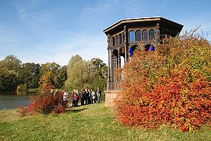 Eine Gruppe steht vor einem großen gusseisernen Pavillon mit bunten Fenstern im oberen Teil. Die Sonne scheint und lässt das rote Herbstlaub der Sträucher um den Pavillon leuchten.