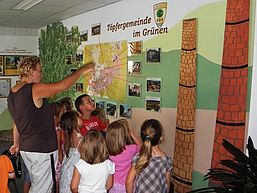 eine Frau zeigt einer Gruppe Kinder etwas an einer mit Karte, Bildern und Zeichnungen gestalteten Wand