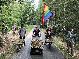 Drei junge Leute mit Schubkarre bzw. kleinem Dumper voller grober Steine. 