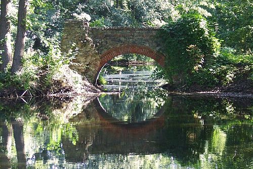 Inmitten eines dschungelartigen Blättergewirr überspannt eine kleine Brücke aus groben Feldsteinen eine Wasserfläche  - und spiegelt sich darin wider.